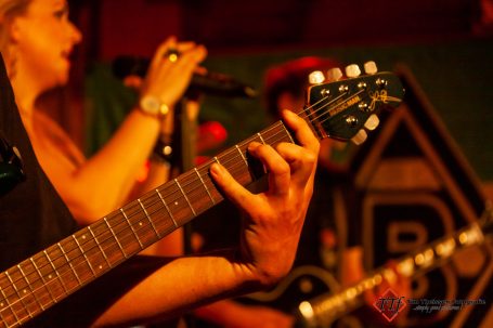 Eine Hand greift eine E-Gitarre, während im Hintergrund Sänger und Musiker auftreten.