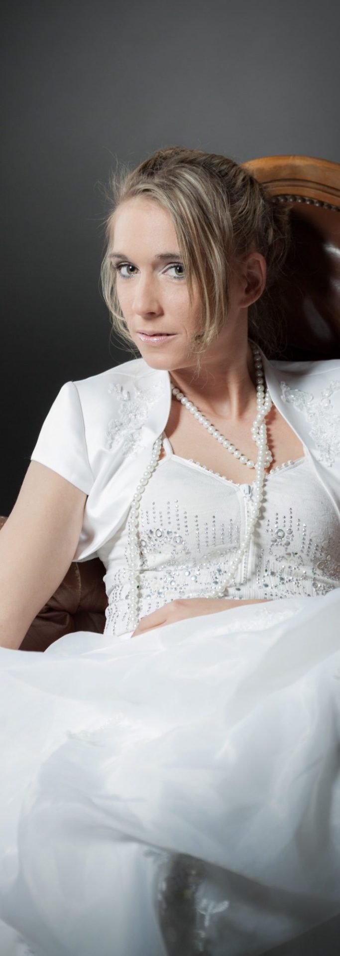 Junge Frau in einem weißen Brautkleid mit einer eleganten Jacke und einer Perlenkette.