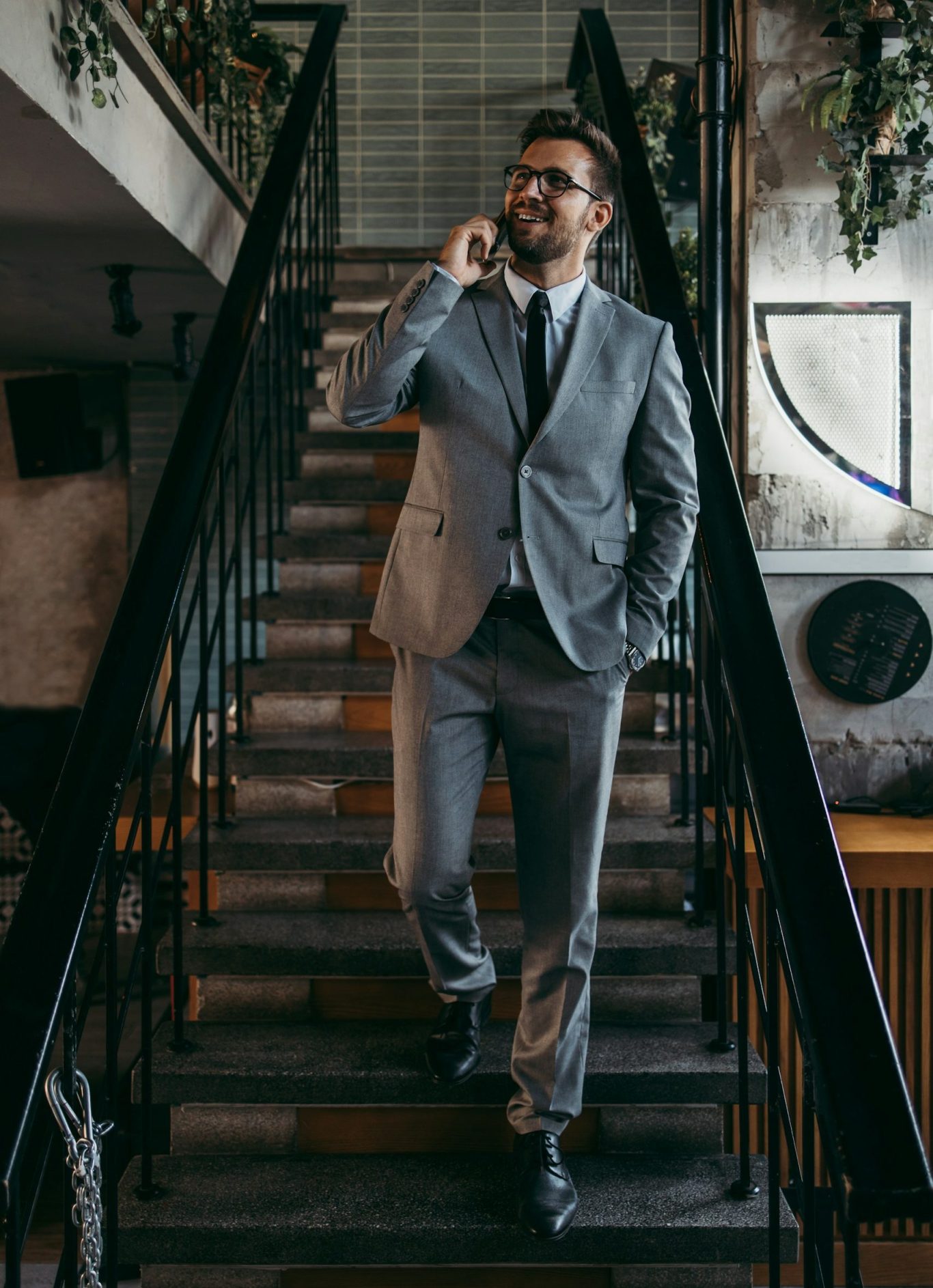 Businessfotografie Mann mit Bart und Brille geht in einem grauen Anzug die Treppe hinunter. Dabei telefoniert er und lächelt.