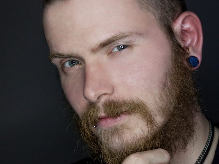 Männlicher Porträt mit Bart, blauen Augen und Ohrstecker, nachdenklicher Ausdruck.