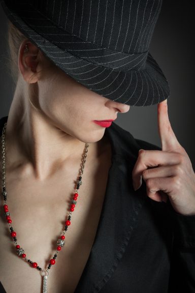 Eine Person mit einem Hut und roten Lippen posiert geheimnisvoll in einem schwarzen Outfit.