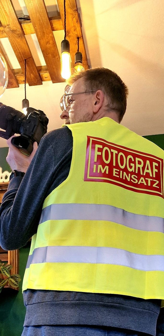 Fotograf im Einsatz Ein Mann in einem gelben Warnweste mit der Aufschrift "FOTOGRAF" hält eine Kamera.