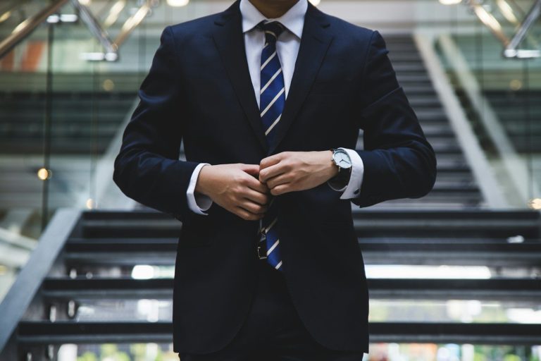 Business Fotos Mann in Anzug mit Krawatte, der sich am Hemdkragen richtet, vor einer Treppe.