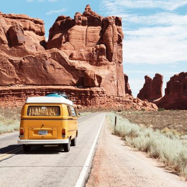 Gelber VW-Bus fährt auf einer Straße neben roten Felsen und einer grünen Landschaft.