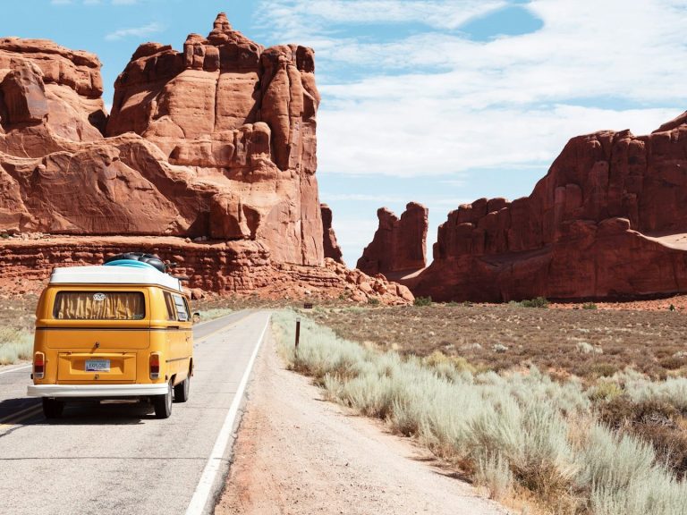 Gelber Volkswagen-Bus fährt auf einer Straße durch eine rotbraune, felsige Landschaft.
