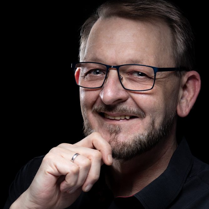 Mann mit Brille lächelt und stützt das Kinn auf die Hand, vor schwarzem Hintergrund.