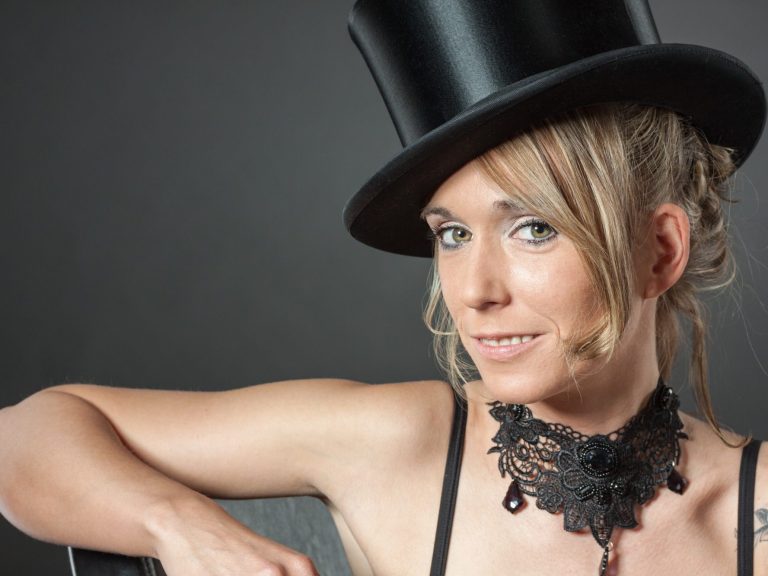 Junge Frau mit zylinderartigem Hut und schwarzem Spitzen-Halsband, lächelt.