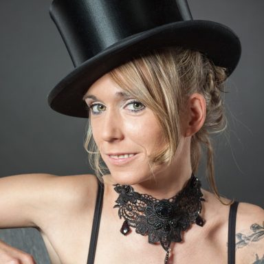 Frau mit einem hohen Zylinderhut und einem eleganten Halsband, lächelt leicht.