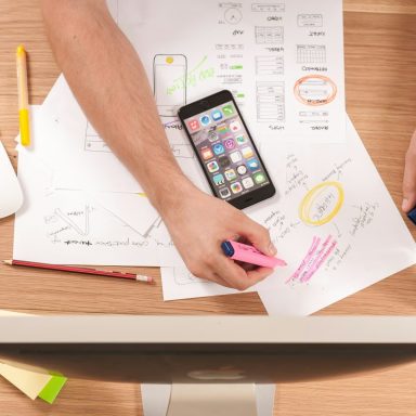 Hände markieren Entwürfe und Notizen auf einem Tisch, neben einem Smartphone und einem Computer.