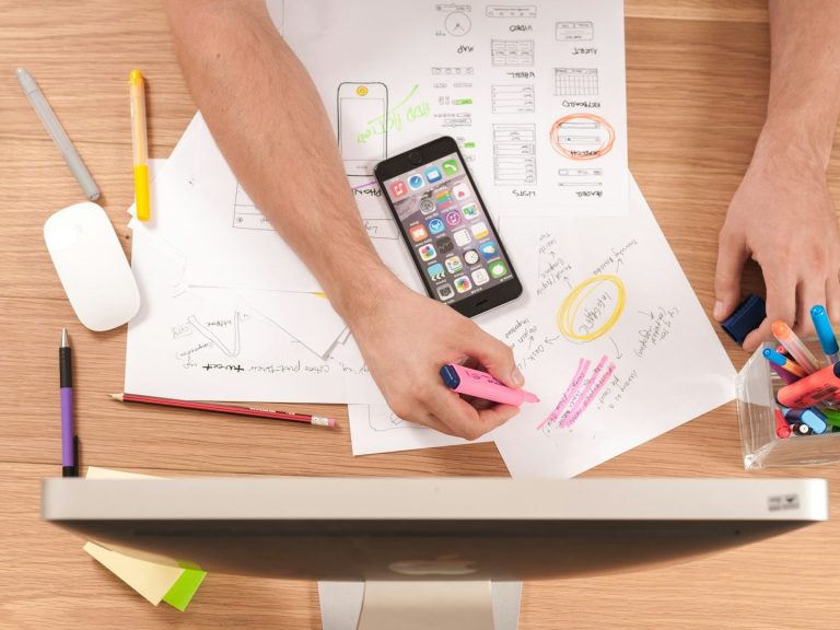 Hände mit Stiften und Notizen neben einem Smartphone auf einem Tisch.