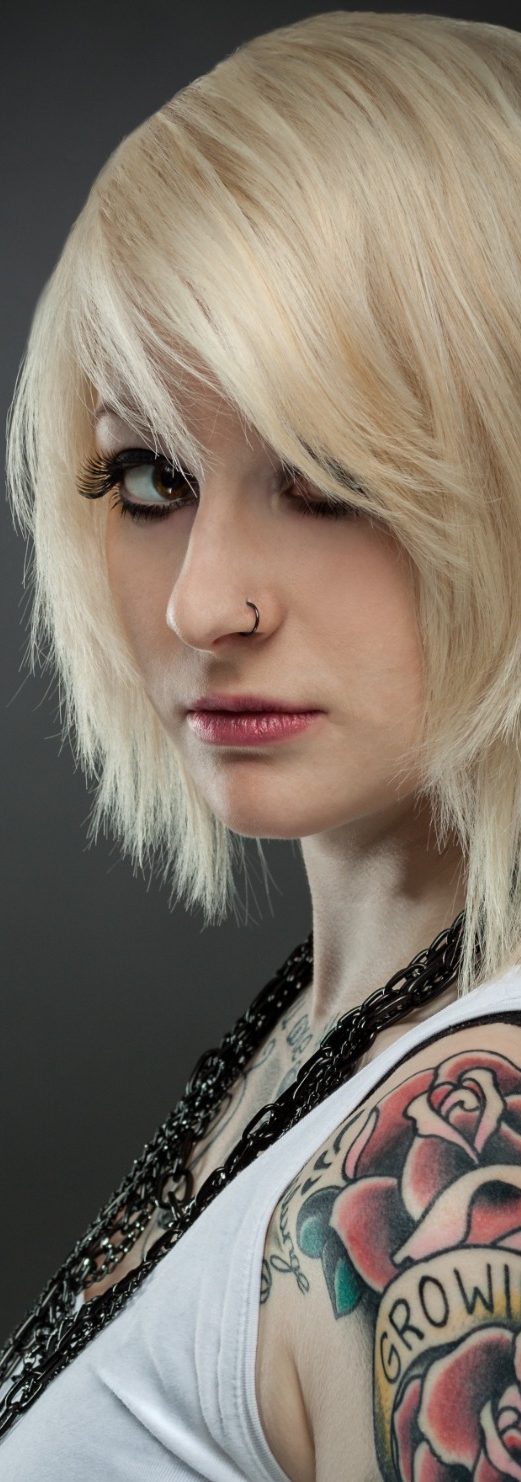 Blonde Frau mit kurzen Haaren und auffälligem Tattoo auf dem Arm, schaut direkt.