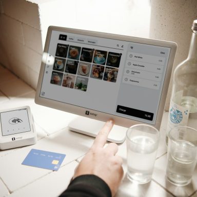 Ein Finger zeigt auf einen Computerbildschirm mit Fotos, daneben ein Glas Wasser und eine Flasche.