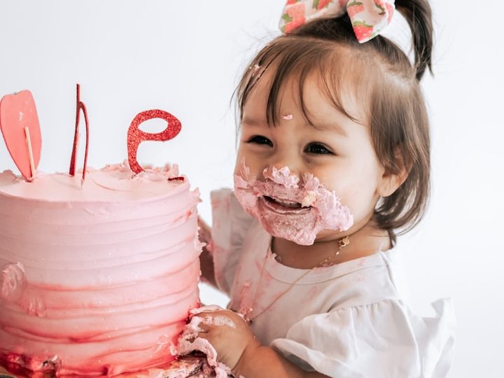Ein kleines Kind mit Sahne im Gesicht steht neben einer rosa Geburtstagstorte.