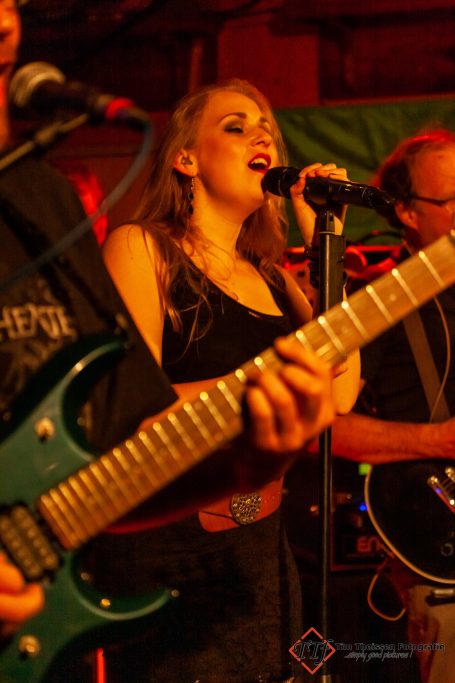Frau mit Mikrofon singt, umgeben von Musikern mit Gitarren in einem lebhaften Umfeld.
