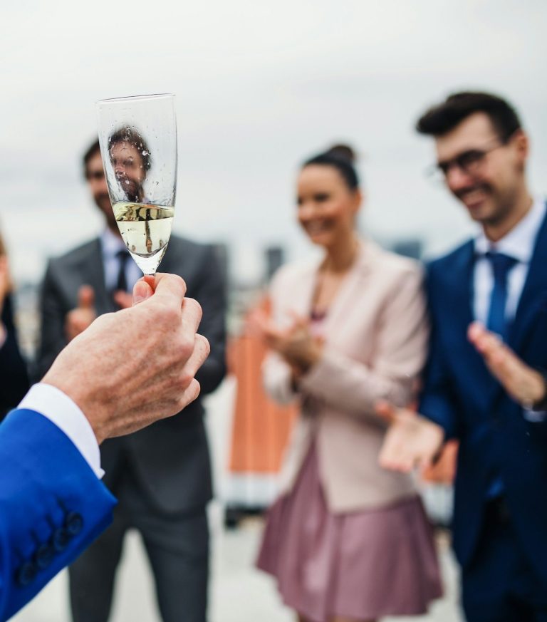 Firmenevent buchen Mann sieht durch ein Sektglas welches von einem Redner gehalten wird in die Gesichter der Leute die zuhören