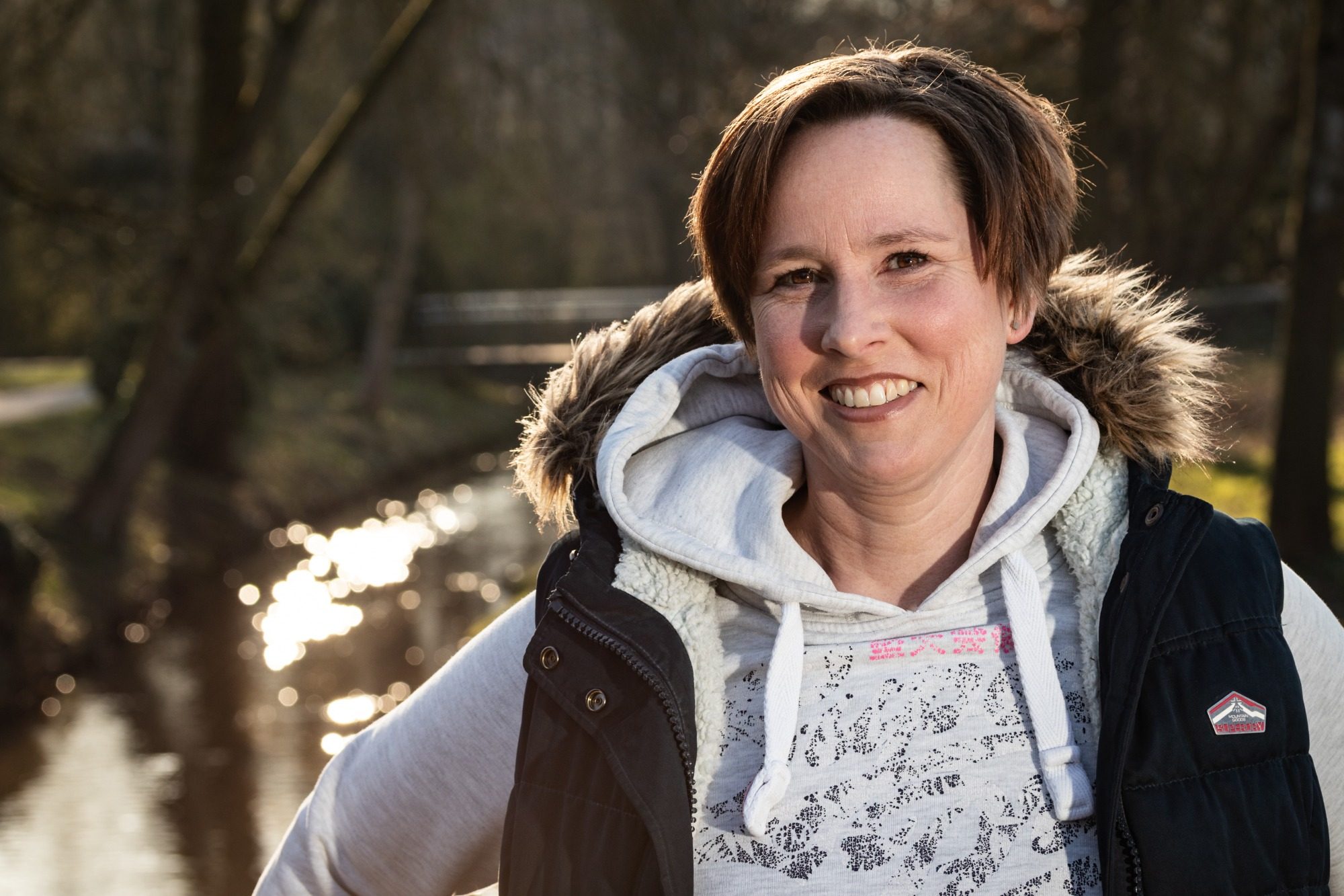 Portraits Frau mit lächelndem Gesicht, trägt eine Jacke, steht am Ufer eines ruhigen Gewässers.