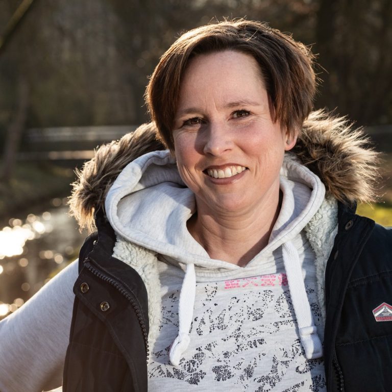 Tanja Lächelnde Frau mit kurzer Frisur, wearing a hoodie und Weste, vor einem Gewässer im Hintergrund.