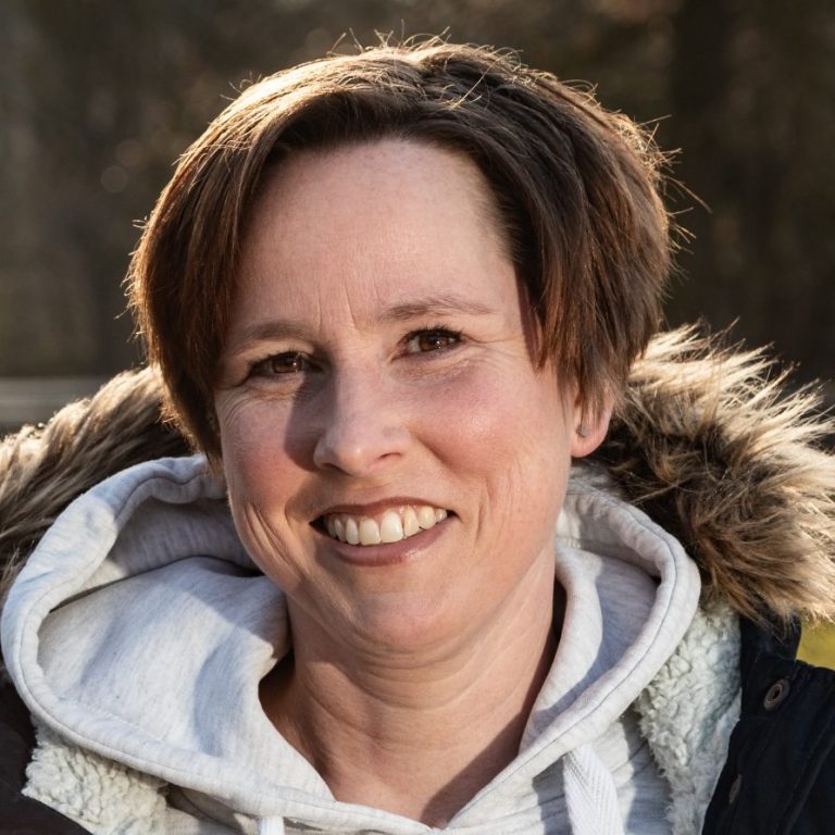 Frau mit lässiger Kleidung und Pelzkragen lächelt vor einer natürlichen Kulisse.
