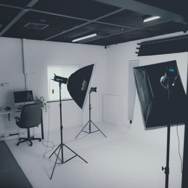 Heller Fotostudio mit Stativ, Softboxen und einem Schreibtisch in der Ecke.