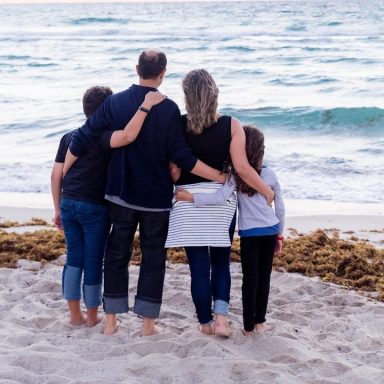 Familie steht am Strand und blickt auf das Meer, umarmt von hinten.