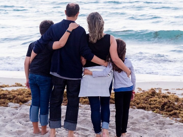 Familie steht am Strand, Blick aufs Meer, Kinder umarmen die Erwachsenen.