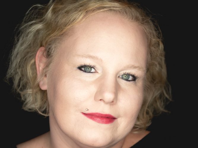 Porträt einer lächelnden Frau mit blonden, lockigen Haaren und rotem Lippenstift.