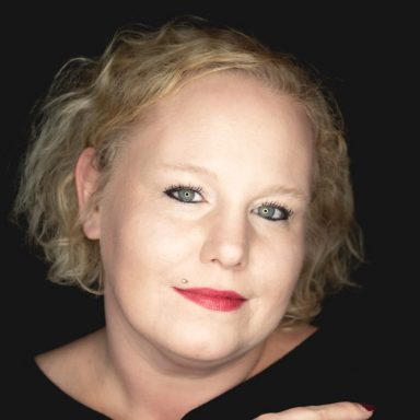Frau mit blonden, lockigen Haaren und rotem Lippenstift, lächelt in die Kamera.