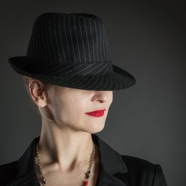 Frau mit schickem Hut, roten Lippen und formeller Kleidung, schaut geheimnisvoll.