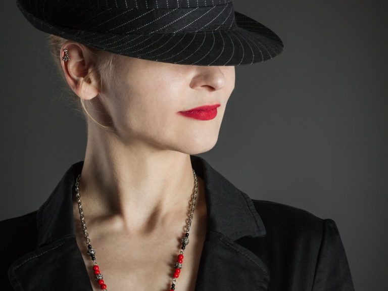 Frau mit Hut, rotem Lippenstift und schwarzem Blazer, trägt eine Halskette.