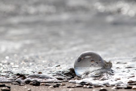 Wassertropfen auf einem sandigen Untergrund, reflektiert Licht und Umgebung.