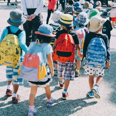 Gruppe von Kindern mit bunten Rucksäcken und Hüten, die in einer Reihe gehen.