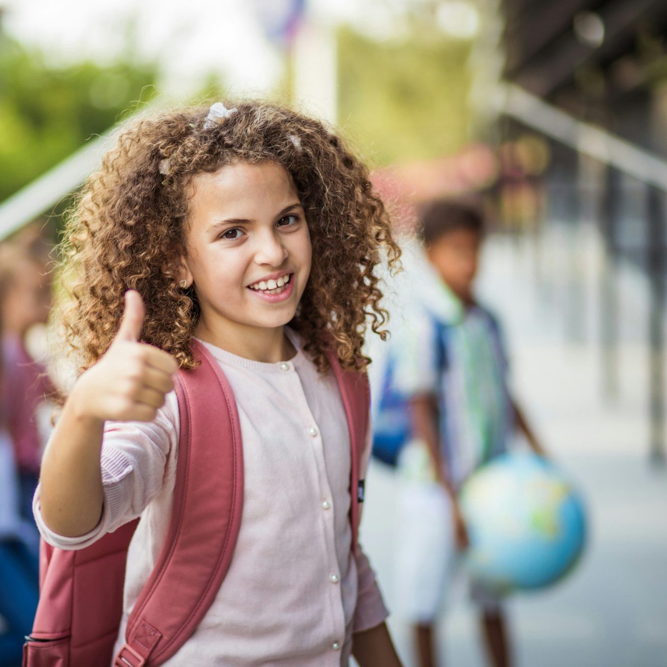Einschulung Mädchen mit lockigen Haaren und eine Schultonister zeigt Daumen hoch
