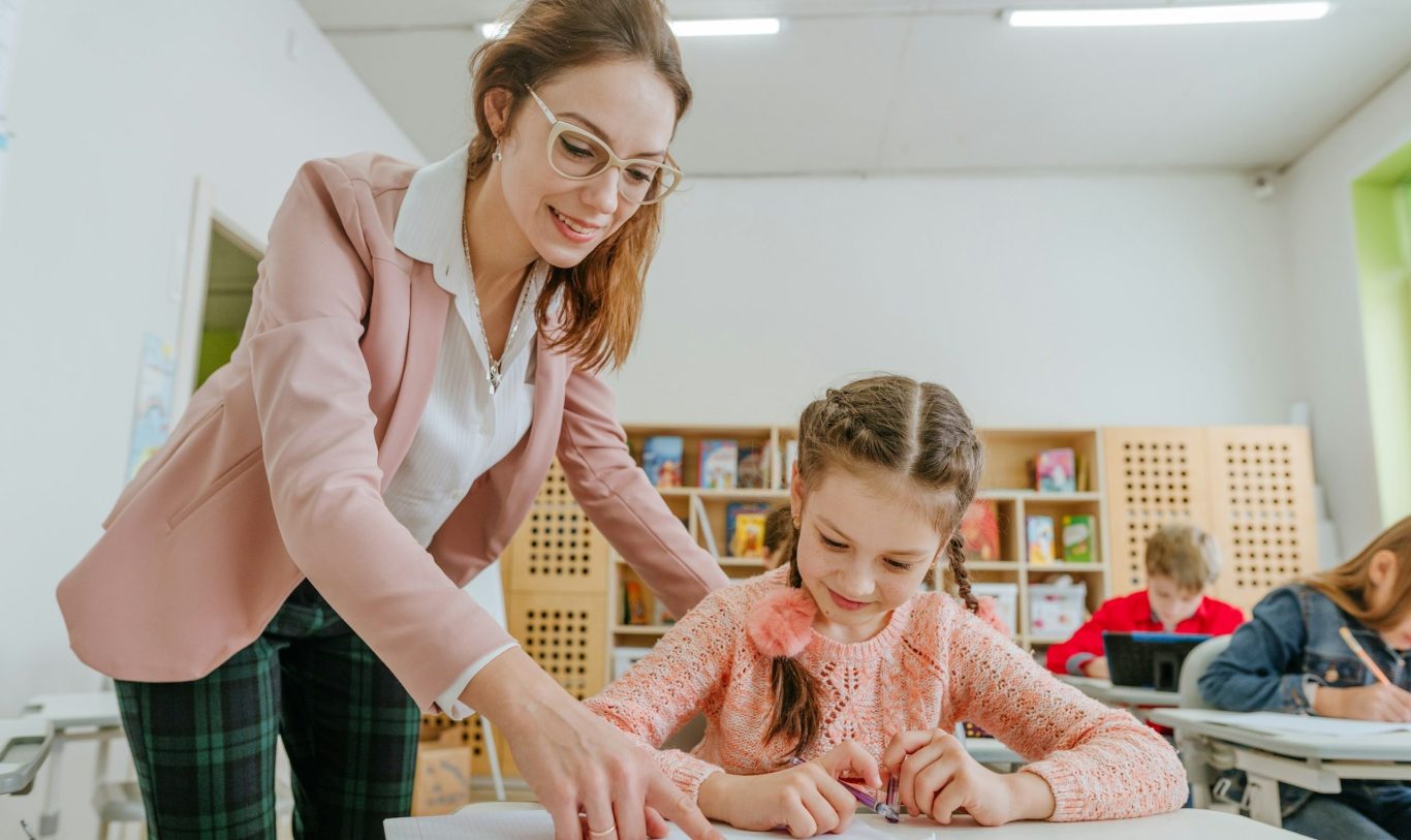 Schulreportage Winw junge Lehrerin mit Brille und langen braunen Harren in einem Rose Sakko gekleidet erklärt einem Mädchen mit Zöpfen etwas. Beide lächeln