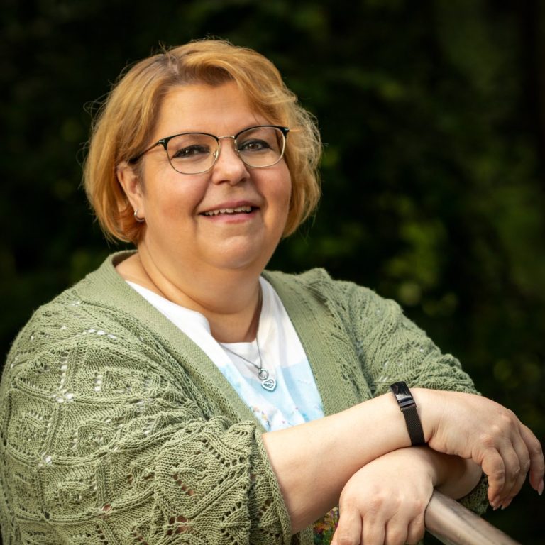 Tanja Lächelnde Frau mit kurzer Frisur, wearing a hoodie und Weste, vor einem Gewässer im Hintergrund.
