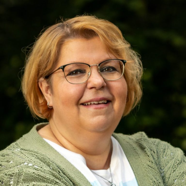 Lächelnde Frau mit blonden Haaren, lässig gekleidet, steht an einem Geländer im Freien.